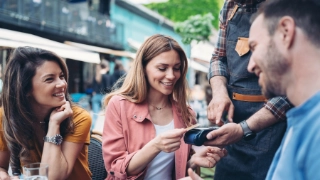 Freunde sitzen draussen beim Kaffee und eine Frau zahlt mit Kreditkarte